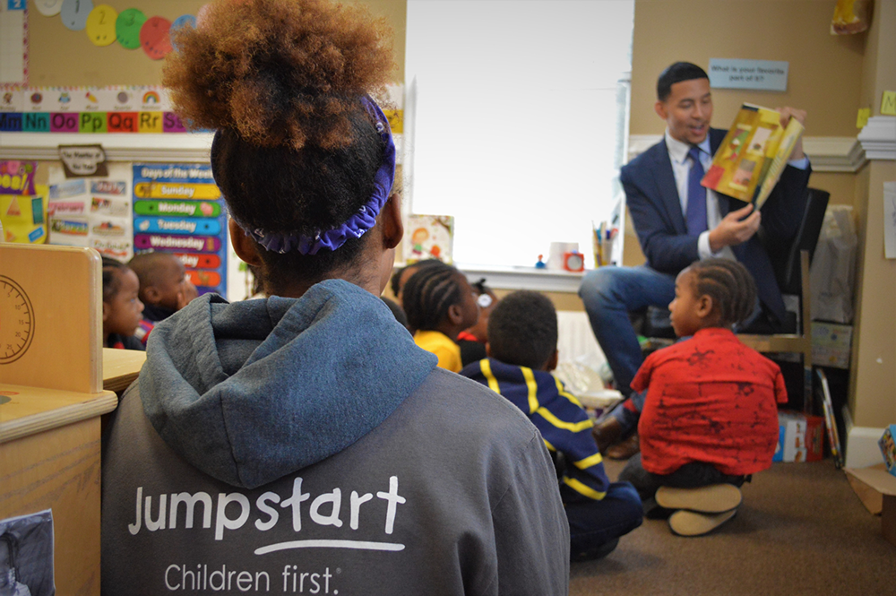 The backside of woman with the Jumpstart logo on her sweatshirt who looks onto a classroom of children and a teacher reading to them.